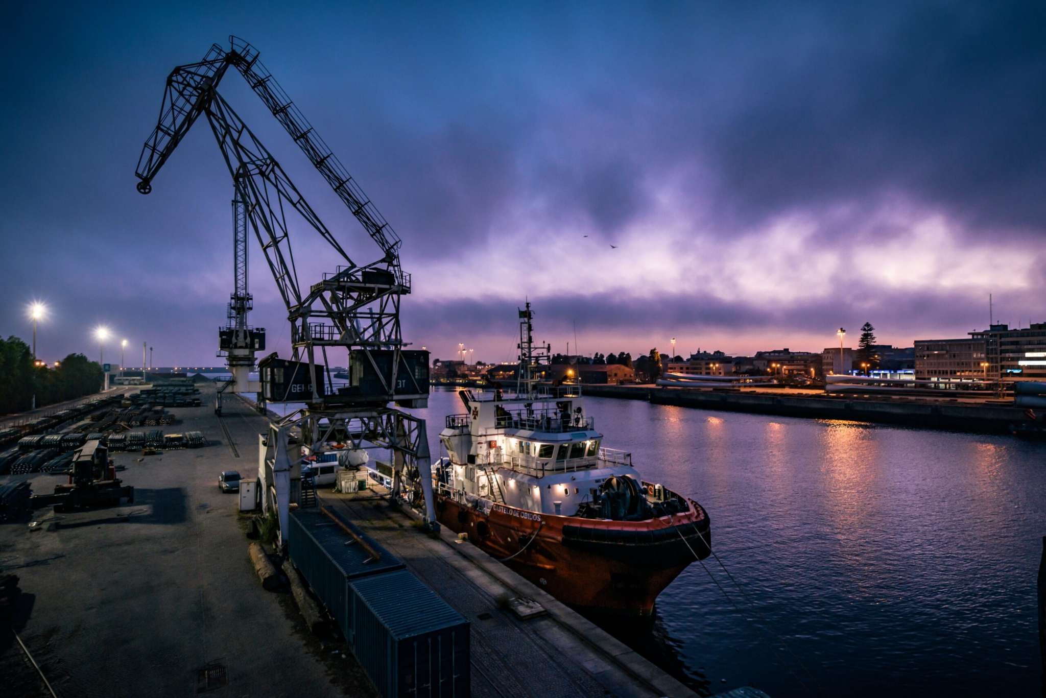 Cargo handling cranes and a tugboat beside a harbour at dusk